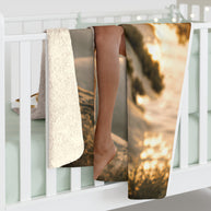 A folded blanket or throw draped over a white crib, showing a printed beach sunset scene with warm golden light, dark tree silhouette, and a cream sherpa-like fleece side. A person's lower legs and bare foot are visible resting on the blanket, and the crib is softly lit in a bedroom setting.