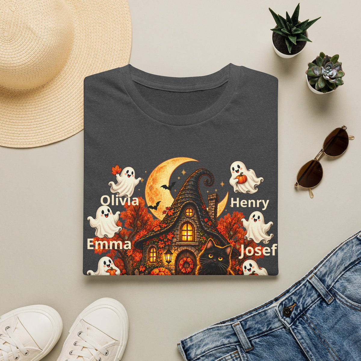A folded dark gray graphic T-shirt is shown in a styled flat lay with a straw hat, small potted succulents, sunglasses, white sneakers, and blue jeans around it. The shirt features a Halloween scene with a haunted house, orange moon, ghosts, a black cat, and the names Olivia, Emma, Henry, Josef, and Joseph printed in white.