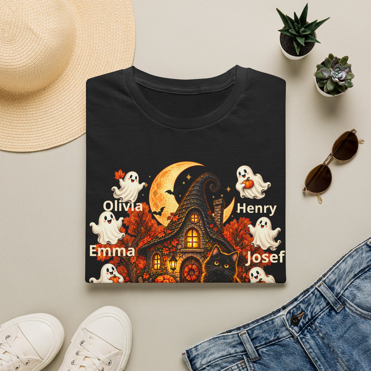 A black short-sleeve T-shirt laid flat on a light beige surface, styled with a straw hat, small potted succulents, sunglasses, white sneakers, and blue jeans. The shirt features a Halloween-themed graphic with a haunted house, orange moon, ghosts, a black cat, and the names Olivia, Emma, Henry, Josef, and Joseph printed in white.