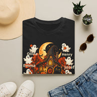 A black short-sleeve T-shirt laid flat on a light beige surface, styled with a straw hat, small potted succulents, sunglasses, white sneakers, and blue jeans. The shirt features a Halloween-themed graphic with a haunted house, orange moon, ghosts, a black cat, and the names Olivia, Emma, Henry, Josef, and Joseph printed in white.