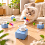 A fluffy cream-colored cat plays with a blue and white laser toy on a wooden floor in a bright living room. The toy consists of four blue cube bases with white bowls, connected by a string with colorful pom-poms and feathers.