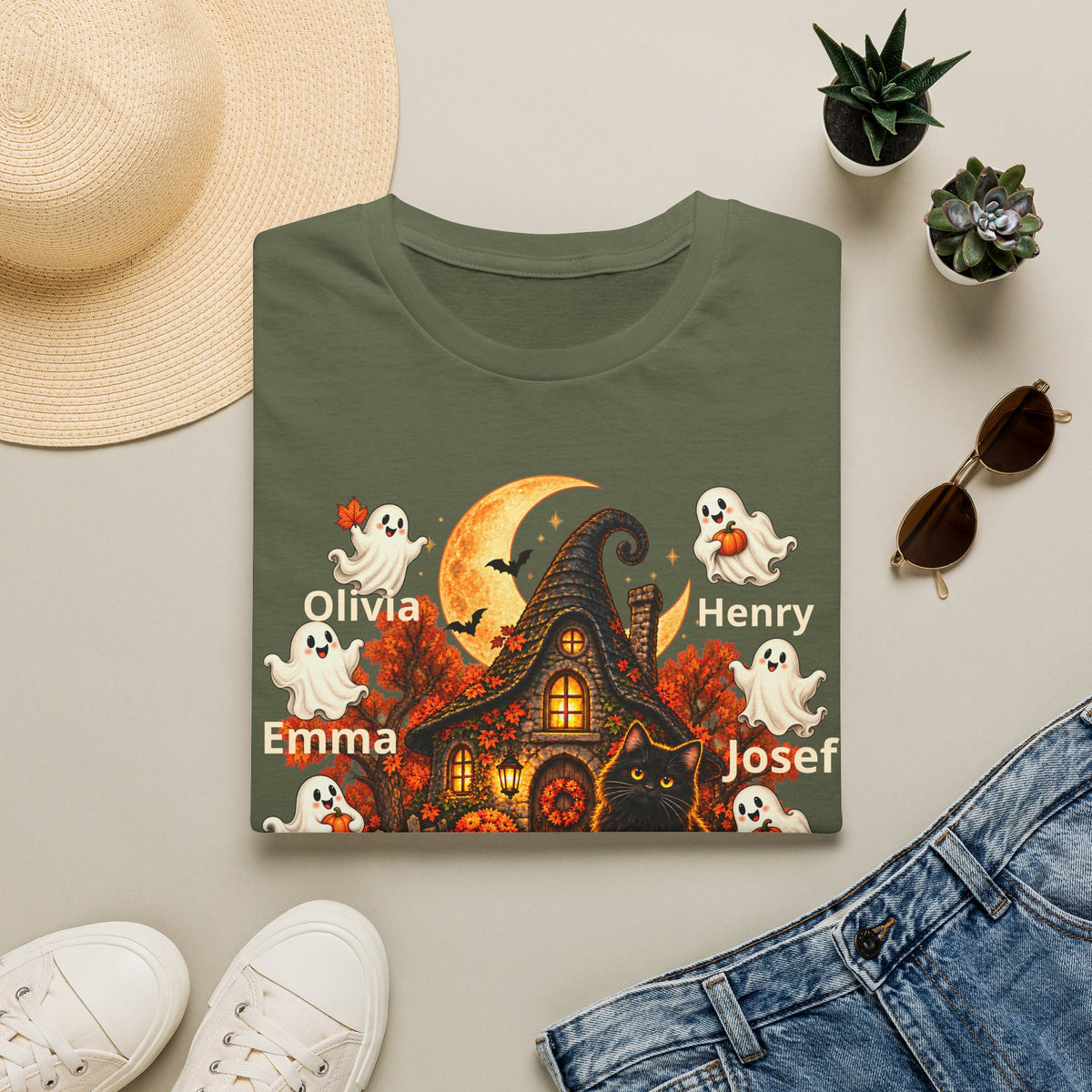 An olive green short-sleeve T-shirt laid flat on a light beige surface, styled with a straw hat, small potted succulents, sunglasses, white sneakers, and blue jeans. The shirt features a Halloween-themed graphic with a haunted house, orange moon, ghosts, bats, a black cat, and the names Olivia, Emma, Henry, and Josef printed in white.