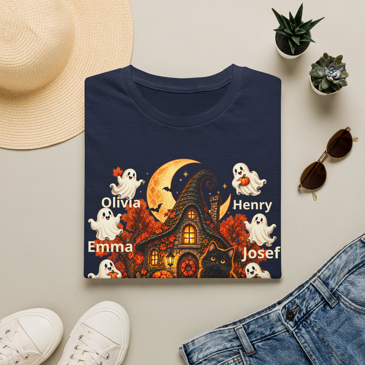 A navy blue folded T-shirt with a colorful Halloween graphic print featuring a haunted house, ghosts, a cat, a crescent moon, and the names Olivia, Emma, Henry, and Josef, styled in a flat lay with a straw hat, sunglasses, jeans, and white sneakers on a light beige background.