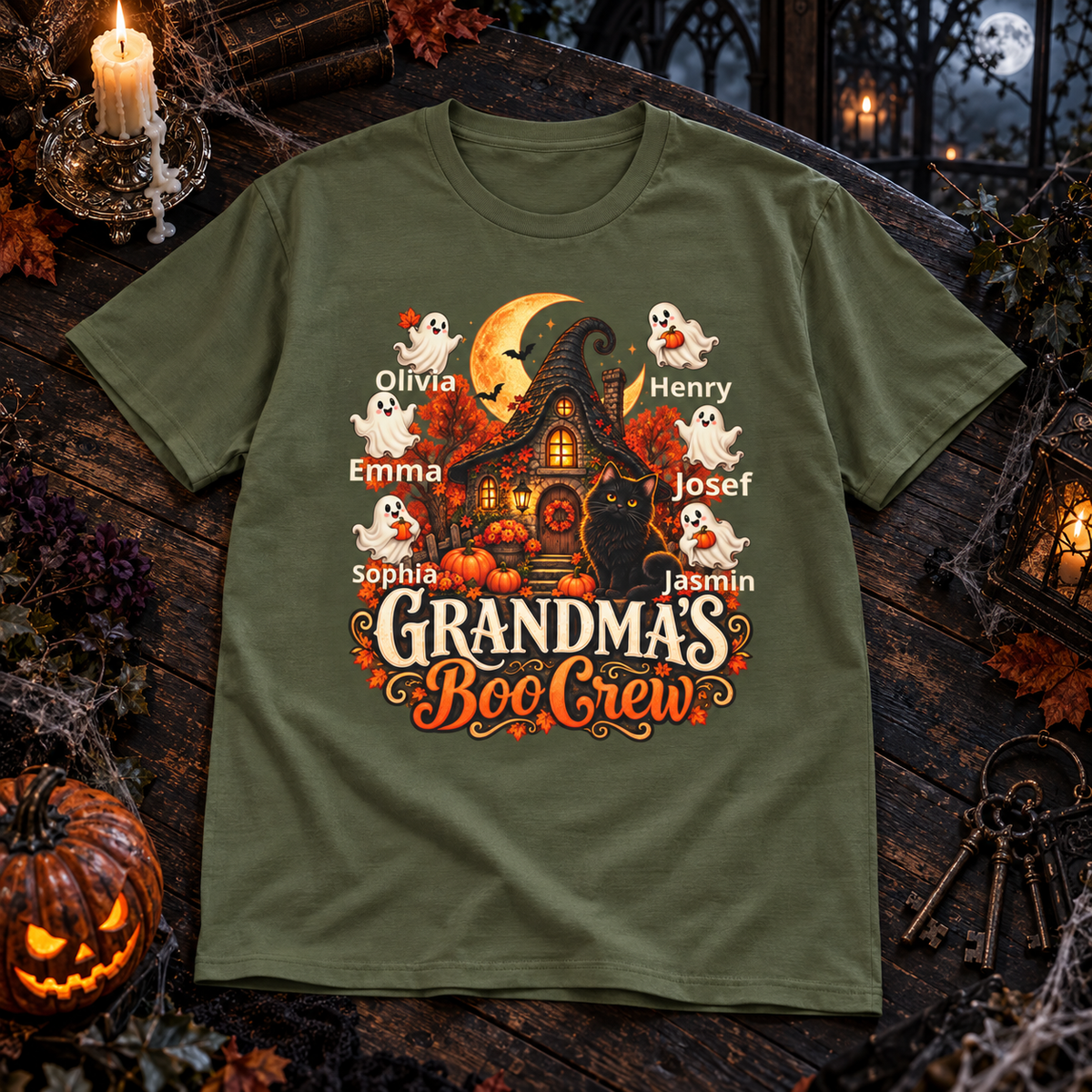 An olive green short-sleeve t-shirt with a detailed Halloween graphic on the front, laid flat on a dark rustic wooden surface surrounded by seasonal props like a jack-o'-lantern, candles, and keys.