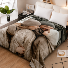 A bed in a styled bedroom featuring a large photographic print of a couple hugging under cozy sweaters and scarves. The bedding includes white pillows and a beige cable-knit throw blanket, set against a warm wood floor and bedside tables.
