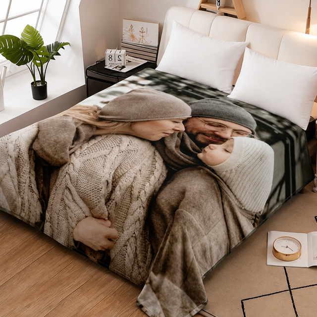 A bed in a styled bedroom featuring a large photographic print of a couple hugging under cozy sweaters and scarves. The bedding includes white pillows and a beige cable-knit throw blanket, set against a warm wood floor and bedside tables.
