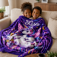 Two children smiling while wrapped in a plush purple blanket featuring a white unicorn, stars, flowers, and the name 'Lisa' in white script, sitting on a beige couch in a living room.