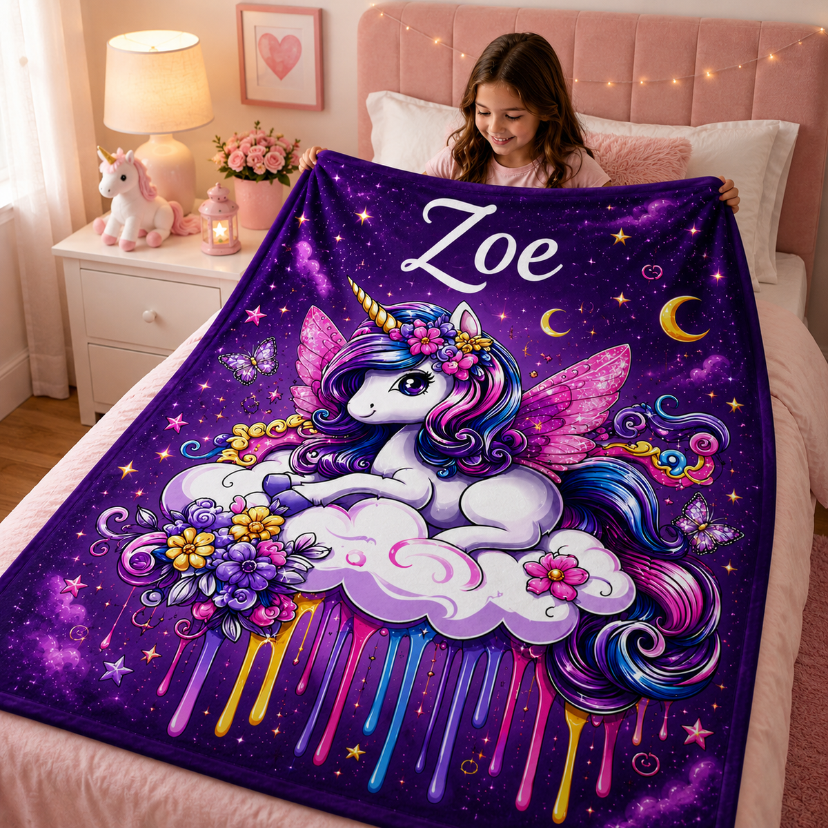 A girl holds up a large personalized blanket on a bed in a cozy bedroom. The blanket features a colorful fantasy unicorn illustration with pink wings, a unicorn horn, flowers, stars, moons, and the name 'Zoe' in white script at the top.