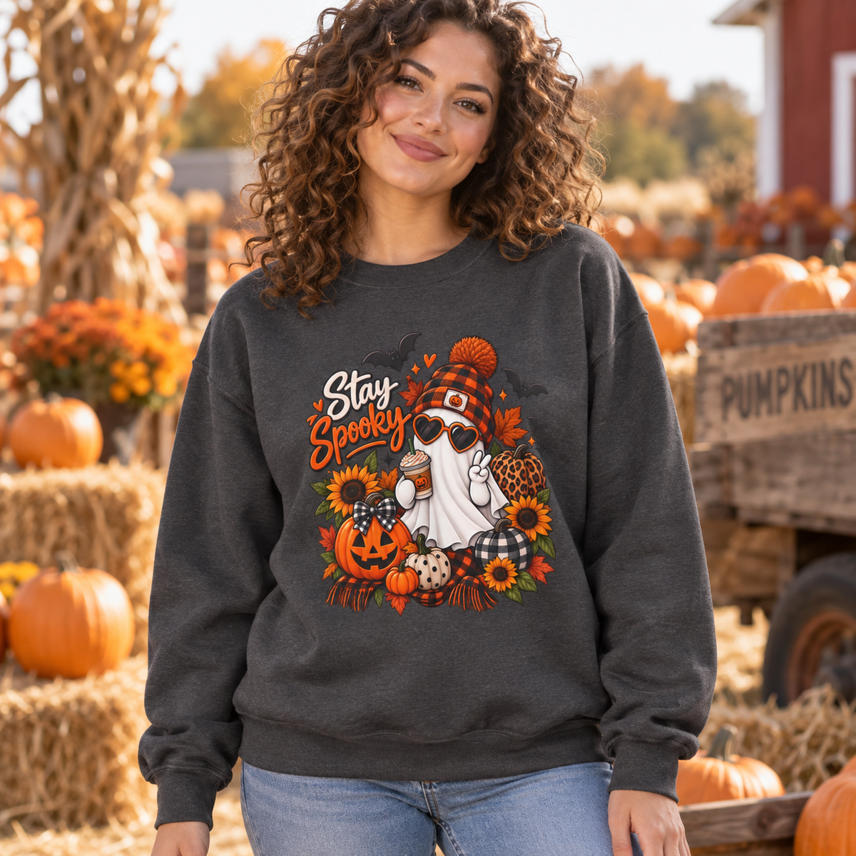 A woman wearing a dark gray crewneck sweatshirt with a 'Stay Spooky' autumn graphic, standing in a pumpkin patch with hay bales and pumpkins in the background.