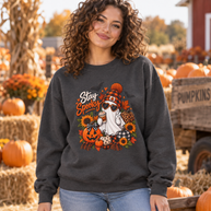 A woman wearing a dark gray crewneck sweatshirt with a 'Stay Spooky' autumn graphic, standing in a pumpkin patch with hay bales and pumpkins in the background.