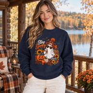 A woman stands on a wooden porch wearing a navy blue sweatshirt with a colorful 'Stay Spooky' graphic featuring a ghost, sunflowers, pumpkins, and autumn leaves. The background shows a scenic lake and autumn forest.