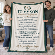 A man and a woman hold up a large cream-colored blanket with a dark green border and a printed poem titled 'TO MY SON' in dark green and brown text. The photo is taken in a living room setting.