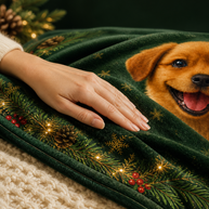 A close-up of a dark green Christmas-themed throw blanket with a printed golden dog face, pinecones, snowflakes, red berries, and warm string lights, draped over a cream knit surface with a hand resting on it.