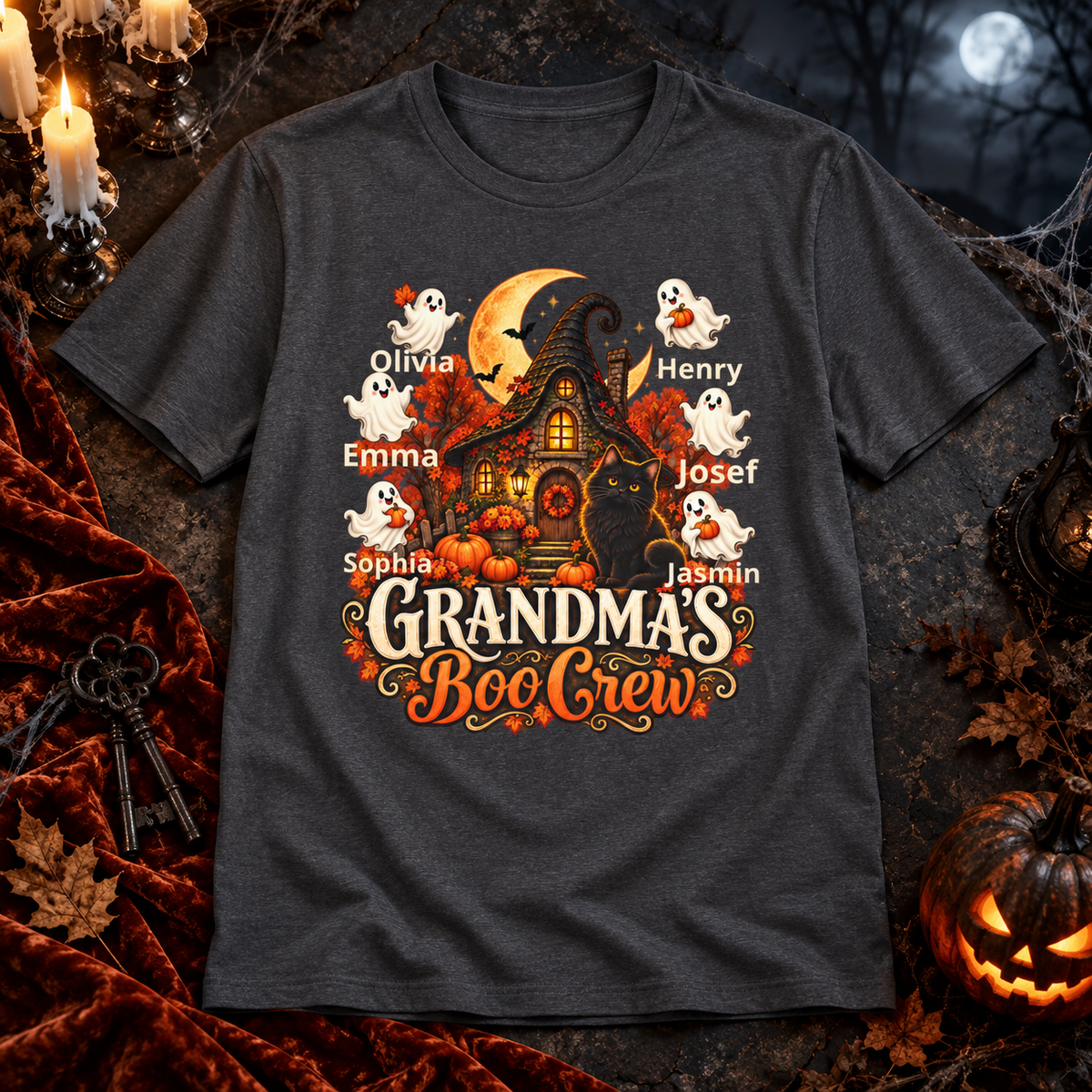 A dark gray graphic t-shirt featuring a Halloween-themed design with a haunted house, pumpkins, ghosts, a black cat, and the text 'Grandma's Boo Crew' with customizable names. The shirt is displayed in a cozy, themed flat-lay setting with autumn props like a carved pumpkin, velvet fabric, and candles.