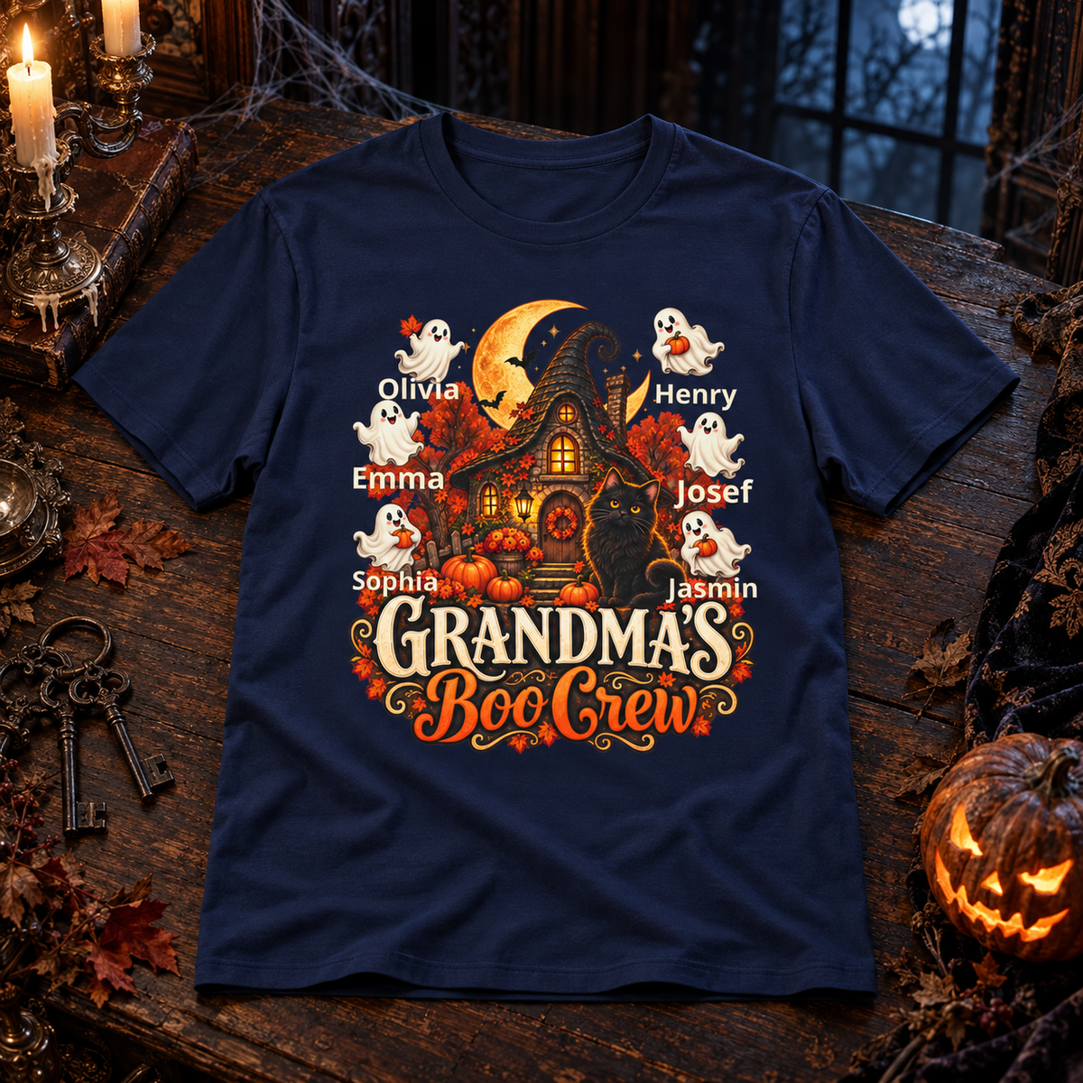 A navy blue short-sleeve graphic T-shirt laid flat on a dark Halloween-themed surface with candles, keys, spiderwebs, autumn leaves, and a jack-o'-lantern. The shirt features a large front print of a haunted house, ghosts, pumpkins, and the text 'Grandma's Boo Crew' with personalized names around the illustration.