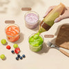 A hand squeezes a green juice into a small glass cup. Other labeled juice cups containing orange carrot juice, purple sweet potato juice, and green kiwi juice are arranged on a beige surface with grapes, berries, a fork, and a wooden board nearby.