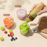 A hand squeezes a green juice into a small glass cup. Other labeled juice cups containing orange carrot juice, purple sweet potato juice, and green kiwi juice are arranged on a beige surface with grapes, berries, a fork, and a wooden board nearby.
